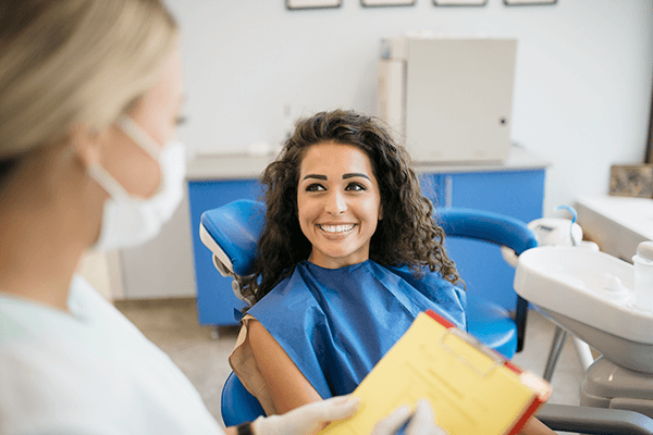 A dental professional in a mask and gloves is consulting a patient in a chair with a clipboard in a clinical office setting.